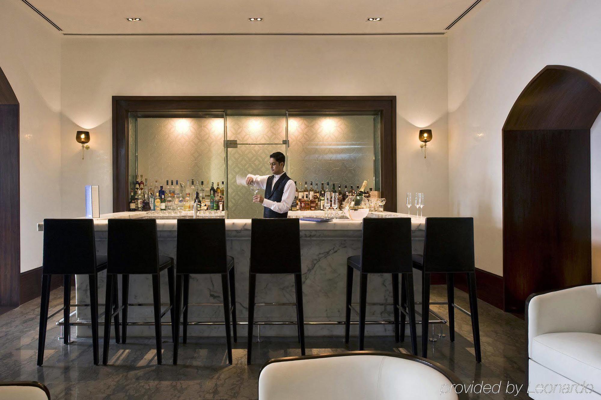 The Taj Mahal Tower, Mumbai Ξενοδοχείο Εστιατόριο φωτογραφία The photo shows a stylish bar area. In the center, a bartender, dressed in a formal outfit, is standing behind a sleek, modern bar counter made of marble. The bar is lined with high chairs for patrons. Behind the bartender, there are shelves filled w